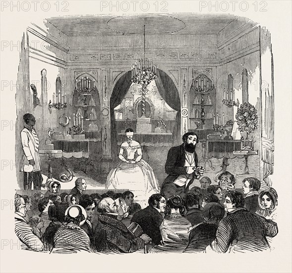 THE SALLE DE ROBIN, PICCADILLY, LONDON, UK. M. Robin leads on Madame blindfolded, and seats her on an ottoman in the centre of the stage, fronting the audience; having done which, he traverses the platform from the stage to the boxes, and gathers from the assembly watches, trinkets, rings, and other articles, and calls upon the supposed clairvoyant to describe them, which she does without hesitation, and with the most perfect exactness., 1851 engraving