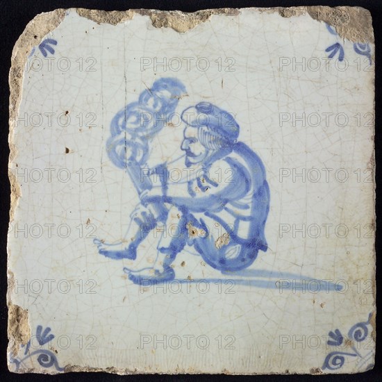 Tile with seated man with beret holding smoking tube or wooden trowel in the hand and or lighting or tapping, corner motif, ox