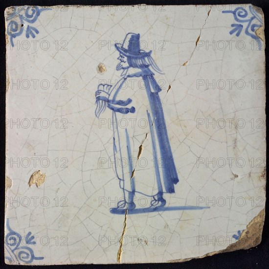 Occupation tile, blue with standing nobleman with handkerchief In hand, long cloak, top hat, corner pattern ox head, wall tile
