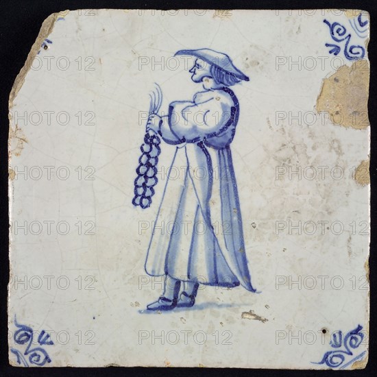 Figure tile, blue with standing figure with long cloak or dress and string of garlic in the hand, corner pattern ox head, wall