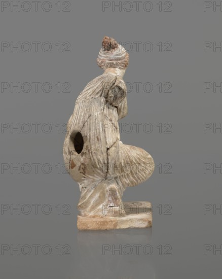 Terracotta figurine of a girl throwing dice. Figurine of a crouching girl throwing dice: weight of the body rests on the left leg, bent at a right angle. The girl wears a long, finely pleated, sleeveless chiton. Hair tied in a bun with a wide headband kredemnos. Earring visible in left ear. Entire figurine rests on a rectangular base. Preservation: missing the right arm; glued in several places head, left arm from 2 fragments, robe, base from 3 fragments. Part of the base filled with clay and plaster. 4th–3rd century BC, Tanagra Greece. Terracotta figurine, height 13 cm, width 9 cm, depth 4.5 cm., A weathered carved figure viewed from the side, showing a textured surface with visible grooves and cracks; the figure has a rounded head with a coiled or knotted element on top and a neck and shoulder area leading into a draped, ribbed garment that folds over a bent limb and forms a rounded, shell-like shape at the front. The sculpture rests on a rectangular base and has a hollowed cavity vi...