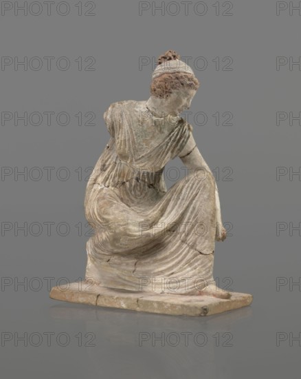 Terracotta figurine of a girl throwing dice. Female figurine of a crouching girl throwing dice; weight of the body rests on the left leg, bent at a right angle. Dressed in a long, finely pleated, sleeveless chiton. Hair tied in a bun with a wide headband kredemnos. Earring visible in the left ear. Entire figurine rests on a rectangular base. Preservation: missing the right arm; figurine glued in several places head; left arm from 2 fragments; robe; base from 3 fragments. Part of the base filled with clay and plaster. 4th–3rd century BC, Tanagra Greece. Terracotta, mold-made figurine; height 13 cm, width 9 cm, depth 4.5 cm., A weathered statue of a seated female figure on a rectangular base, shown in a three-quarter view; the figure wears a draped garment with detailed folds and pleats, her head turned to one side with hair gathered in a bun, and one arm resting on her raised knee while the other arm is bent; the surface shows a worn, textured appearance with areas of flaking and cracki...
