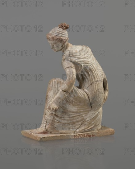 Terracotta figurine of a girl throwing dice. Female figurine in a long, finely pleated sleeveless chiton, hair in a bun tied with a wide headband kredemnos, earring in left ear. Crouching posture with body weight on the left leg bent at a right angle; entire figurine on a rectangular base. Mold-made, Tanagran style. Preservation: missing right arm; glued in several places head; left arm from 2 fragments; robe; base from 3 fragments; part of the base filled with clay and plaster. Date: 4th–3rd century BC; origin: Tanagra Greece. Material: terracotta. Dimensions: height 13 cm, width 9 cm, depth 4.5 cm., A small weathered statue of a seated figure on a rectangular base against a grey background, showing a person with hair gathered in a knotted topknot and wearing a draped garment with visible folds; the figure is bent forward with one arm reaching down toward the knee and the other arm resting near the body, and the surface shows aged, cracked areas and worn paint in tones of off-white, b...