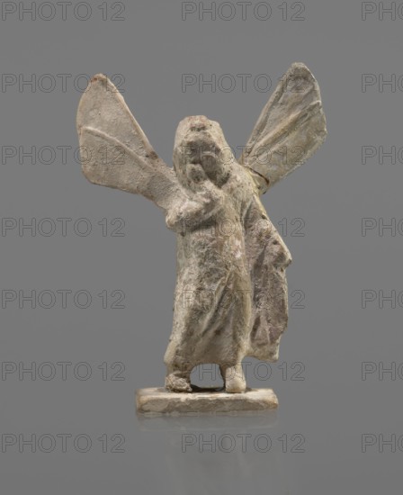 Terracotta figurine of Psyche, Young Psyche with butterfly wings, wrapped in a chlamys. Right hand rests on the chest, left hand rests along the body. Long hair falling on the shoulders is arranged in curls. Head turned to the left. Figurine placed on a rectangular base. 332–30 BC; Hellenistic period. Tanagra Greece. Figurine, height 10.8 cm, width 7.5 cm, depth 3 cm., A small sculpted figure with a textured surface standing on a rectangular base, showing a long draped garment and a flowing cloak, with two large wing-like extensions behind the shoulders; the surface displays rough, worn details and varied tones of beige, off-white, pale gray, and hints of muted brown and faint pinkish areas, all set against a plain medium-gray background.