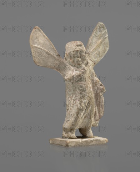 Terracotta figurine of Psyche, Young Psyche with butterfly wings, wrapped in a chlamys. Right hand rests on the chest, left hand rests along the body. Long hair falling on the shoulders arranged in curls. Head turned to the left. Figurine placed on a rectangular base. 332–30 BC; Hellenistic period. Origin: Tanagra Greece. Figurine. Height 10.8 cm, width 7.5 cm, depth 3 cm., A weathered small statue of a standing figure with a rough textured surface, shown from the back, with a pair of large wing-like extensions rising upward from the shoulders; the figure has a short, rounded head and loosely draped clothing that hangs to the base, creating folds and irregular contours. The object rests on a rectangular plinth with a slightly rounded top edge and a worn appearance. The visible colors are muted beige and off-white tones with areas of pale pink and faint gray-green patina, and small darker brownish spots. The background is a uniform medium gray.