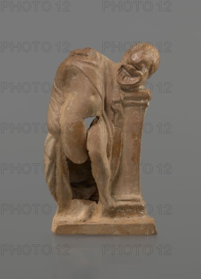 Terracotta figurine of a youth with a terracotta mask; creator, ca. 332–30 BC, Hellenistic period 4th century BC; Myrina; figurine; height 12.3 cm., A small sculptural figure in shades of brown and tan is shown against a gray background, depicting a hunched person leaning on a rectangular pedestal with a rounded head turned to the side and a face with pronounced features, draped in a loose garment that reveals bent legs and bare feet, with areas of lighter and darker brown patina and some worn or chipped spots on the surface.