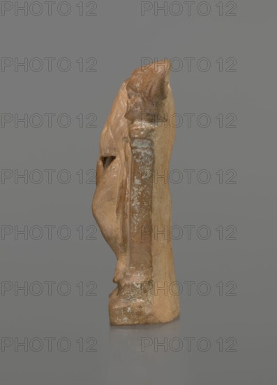 Terracotta figurine of a youth with a terracotta mask, Hellenistic period 4th–1st century BC, ca. 3rd–1st century BC; Myrina; terracotta; figurine; height 12.3 cm., A narrow vertical sculpture viewed from the side showing the profile of a draped figure leaning against a column-like support, with visible surface wear and patches of lighter discoloration; colors visible include shades of brown, tan, beige, and small areas of pale off-white against a neutral gray background.