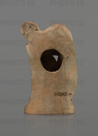 Terracotta figurine of a youth with a terracotta mask; maker, Hellenistic period, circa 332–30 BC 4th century BC; Myrina; figurine; height 12.3 cm; dated ca. 1823–1897 for Hoffmann removed., A roughly shaped vertical object with an irregular surface and a large circular hole through its upper middle, showing an inner dark cavity; the object is primarily tan and light brown with areas of darker brown and subtle grayish tones, and it sits on a reflective gray surface against a smooth medium gray background.