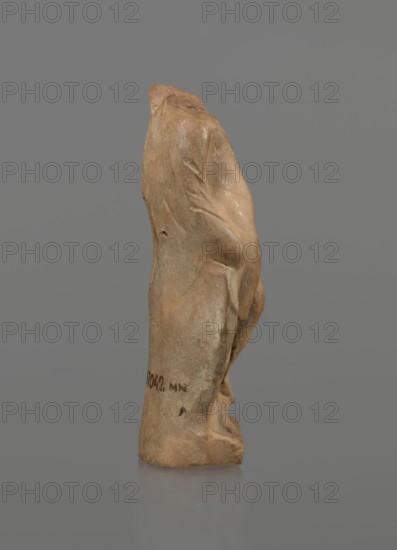 Terracotta figurine of a youth with a terracotta mask, Hellenistic period 4th–1st century BC, ca. 332–30 BC; Myrina; terracotta; figurine; height 12.3 cm; maker, Hoffmann, Henri ca. 1823–1897 mentioned., A small, upright carved object with an irregular, elongated shape appearing to be made of a brownish-beige material with lighter and darker patches, shown against a plain gray background; the surface shows smooth and rough areas with subtle folds and rounded contours suggesting drapery-like forms, and a few small indentations and worn spots; the base rests on a reflective surface that mirrors a muted version of the object’s brown and beige tones.