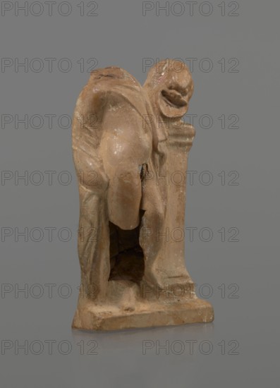 Terracotta figurine of a youth with a terracotta mask; maker, Hellenistic period 4th–1st century BC, ca. 332–30 BC; Myrina; figurine, height 12.3 cm; attributed ca. 4th century BC., A small sculpted figure made of a brownish-beige material stands against a gray background, showing a draped form with rounded and folded surfaces and a hollowed area near its base; the sculpture has smoother and rougher textured areas, with some lighter and darker patches of beige and pale tan, and a few subtle greenish and whitish spots; the base is rectangular and rests on a reflective surface that creates a faint muted reflection.