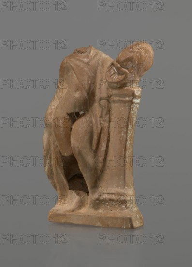 Terracotta figurine of a youth with a terracotta mask, ca. 332–30 BC; Hellenistic period 4th century BC; Myrina; figurine; height 12.3 cm., A weathered brownish clay sculpture of a crouching human figure leaning against a rectangular pillar, showing a rounded head tilted to the side and draped garments that form folds over the torso and legs, with visible surface wear and lighter patches of whitish residue, set against a neutral gray background and resting on a flat base with a subtle reflection.