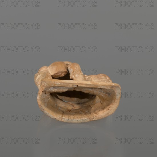 Terracotta figurine of a youth with a terracotta mask, attributed to Hoffmann, Henri ca. 1823–1897 - 30 BC; Hellenistic period 4th century BC, Myrina; figurine, height 12.3 cm, A beige-brown clay object with an irregular hollow center rests on a smooth gray surface against a muted gray background; the object shows rough, uneven edges, patches of lighter and darker brown, and small chips revealing a slightly paler interior, with a few glossy spots and subtle shadowing beneath it.