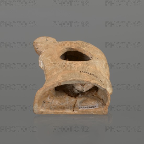 Terracotta figurine of a youth with a terracotta mask;, Hellenistic period, ca. 4th–3rd century BC 30 BC; Myrina; figurine, height 12.3 cm., A single worn object resembling a hollowed vessel with a rounded top and an open cavity is shown against a plain gray background, the object displaying a range of tan, beige, light brown, and ochre tones with patches of darker brown in recessed areas and lighter cream-colored spots along edges; the surface appears textured and irregular with visible cracks and chips, and a small group of darker interior shadows is visible inside the cavity.