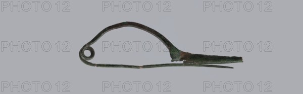 Arched fibula. Fibula with an arched bow; the pin emerges from a double-coiled spring. The sheath is in the form of a half-cylinder, cut to accommodate the pin. Material: bronze. Condition: good, greenish corrosion on the surface. Date: 8th–7th century BC 4th quarter of the 8th – mid-7th century BC. Type: jewelry, fibula. Place of origin: Italy . Maker: unknown., A single elongated metal object with a curved upper arm forming an arch and a coiled circular loop at one end, a straight lower arm extending beneath the arch, and a tapered pointed section at the opposite end; the surface shows uneven texture and areas of corrosion with visible rough patches and flaking, and the object displays a mix of dark green, brown, and muted metallic tones against a plain light gray background.