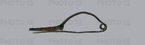 Arched fibula, Goluchów Greater Poland Voivodeship. Fibula with an arched bow. The pin emerges from a double-coiled spring. The sheath is in the form of a half-cylinder, cut to accommodate the pin. Condition: good, greenish corrosion on the surface. Date: 7th–6th century BC / 4th quarter of the 8th – mid-7th century BC Raczynska. Place of origin: Italy . Jewelry, bronze fibula., A narrow metal object with a curved arching top and a coiled circular end, lying horizontally against a plain gray background; the metal shows dark green and brown patina with spots of lighter green corrosion and hints of orange discoloration, and the lower edge includes a thin flat extension that tapers to a point.