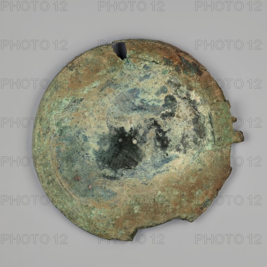 Mirror cover, The cover, with an edge bent inwards at a right angle, is decorated with concentric circles running on both sides at the edge and on the edge. On the outside, a fragment of a hinge with the heads of two rivets is preserved. 425–375 BC, Corinth; mirror cover, height 15.3 cm., A roughly circular, weathered metal object with irregular edges and a few missing fragments, showing mottled patches of green, brown, blue, black, and beige across its surface; there is a darker, irregular stain near the center with two small lighter spots within it, and a small protruding tab on one side.