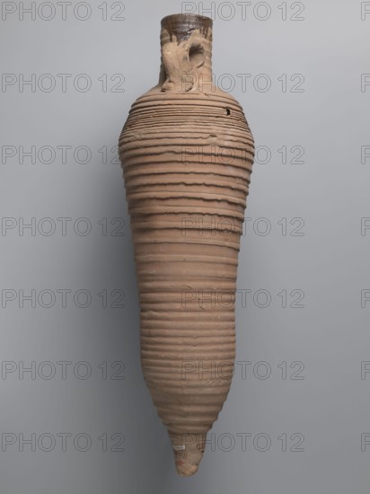 Amphora, unknown Egyptian workshop; wine transport vessel with a strongly elongated belly, narrowed in the middle. Roller-shaped vices, approximately oval in cross-section, are attached to the central section of the neck with a straight rim and connect to the upper part of the convex, steep shoulders. Conical base. Surface brown and even, covered with grooves of varying density and profile. Interior impregnated with a black resinous substance, with streaks visible in the upper neck. Clay brown, alluvial Naqlun AC1A ceramic body type. Inscription in red paint on the upper part of the handles: ICX. Hollowed-out hole ~0.5 cm diameter on the lower part of the handles below the inscription. 2nd half of the 4th–5th century; findspot Edfu. Vessel type: amphora. Dimensions: height 63.6 cm; rim diameter 6.8 cm; body diameter 19.3 cm., An elongated clay vessel with a narrow neck and a pointed base hangs against a pale gray background; the vessel has a ribbed texture formed by a series of horizon...