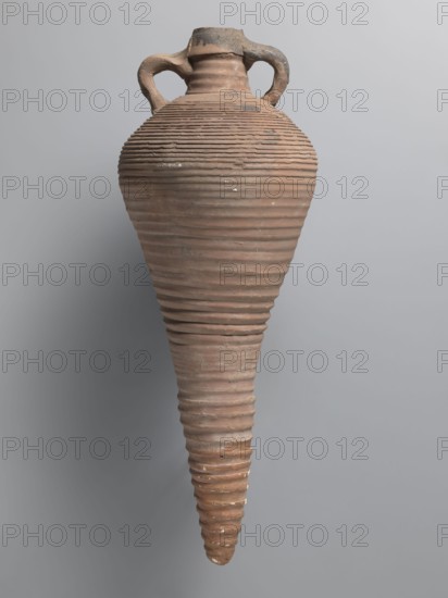 Amphora, unknown Egyptian workshop; storage/transport vessel with a bulging belly, fusiform in the lower part below the point of greatest diameter. Tulip-shaped rim with a chipped finial set on a cylindrical neck. Loop handles attached to the neck and shoulders. Entire brown, even surface covered with dense fluting. Interior impregnated with black resin, with streaks visible on the outside. Conical foot missing; supplemented and reconstructed with plaster. Nile clay; Naqlun AC1A type; 7th–early 8th century; height 45+ cm preserved; lip diameter 5.2 cm; body diameter 21.4 cm., An elongated, ridged amphora-shaped ceramic vessel with a narrow pointed base and a wider rounded shoulder, featuring two looped handles near the short neck; the surface shows pronounced horizontal grooves and irregular worn spots, with visible chips and rough textures. The colors visible are various shades of brown and tan on the vessel, with some darker grayish-brown areas near the rim and handles, and a neutral...