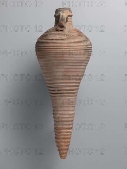 Amphora, unknown Egyptian workshop; amphorae, Egyptian amphorae, Egyptian ceramics, storage vessels, Coptic vessels, amphorae; Nile clay. Transport vessel with a bulging belly, fusiform in the lower part below the point of greatest diameter. Tulip-shaped rim with a chipped finial, set on a cylindrical neck. Loop handles glued to the neck and shoulders. Entire brown, even surface covered with dense fluting. Interior impregnated with black resin, streaks visible on the outside. Conical foot missing; supplemented and reconstructed from plaster. Nile pottery mass Naqlun AC1A, 7th–early 8th century, Edfu; vessel, amphora; height 45+ cm preserved, lip diameter 5.2 cm, body diameter 21.4 cm., A single tall, narrow ceramic vessel with a tapered, conical lower body and a rounded upper body, showing a series of horizontal ridges encircling its surface; the rim at the top is irregular and broken with a small attached fragment. The object displays a weathered, textured surface with visible chips a...