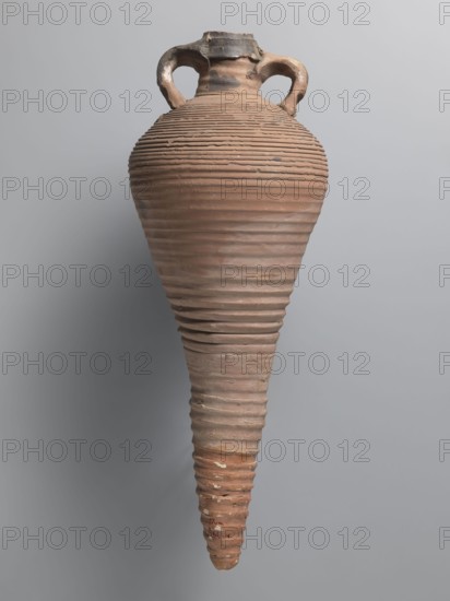 Amphora, unknown Egyptian workshop; amphorae, Egyptian amphorae, Egyptian ceramics, storage vessels, Coptic vessels, amphorae, Nile clay. Transport vessel with a bulging belly, fusiform in the lower part below the point of greatest diameter. Tulip-shaped rim with a chipped finial, set on a cylindrical neck. Loop handles glued to the neck and shoulders. Entire brown, even surface covered with dense fluting. Interior impregnated with black resin, streaks visible on the outside. Conical foot missing; supplemented and reconstructed from plaster. Nile pottery mass Naqlun AC1A, 7th–early 8th century, find from Edfu. Vessel amphora. Height 45+ cm preserved; lip diameter 5.2 cm; body diameter 21.4 cm., An upright, elongated ceramic vessel with a narrow pointed base and a rounded shoulder, featuring a pair of looped handles near the short neck; the surface shows closely spaced horizontal ridges and grooves running around the body, with areas of uneven texture and small chips, and visible color ...