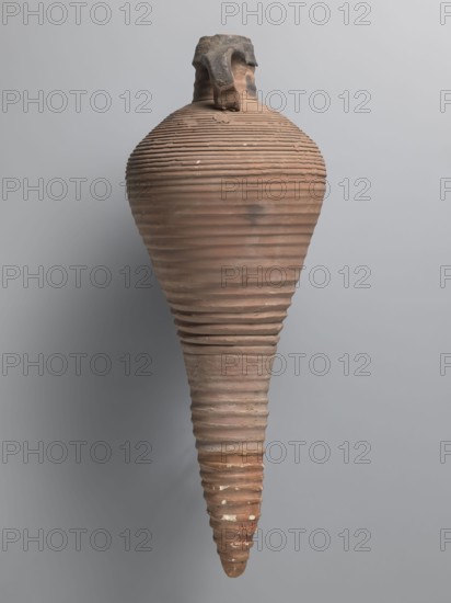 Amphora, unknown Egyptian workshop; transport vessel with a bulging belly, fusiform in the lower part below the point of greatest diameter. Tulip-shaped rim with a chipped finial, set on a cylindrical neck. Loop handles attached to the neck and shoulders. Entire brown, even surface covered with dense fluting. Interior impregnated with black resin, streaks visible on the outside. Conical foot missing; supplemented and reconstructed from plaster. Nile clay, Nile pottery mass Naqlun AC1A, AE/ type amphora, Coptic vessel, 7th–early 8th century, Edfu; vessel amphora, height 45+ cm preserved, lip diameter 5.2 cm, body diameter 21.4 cm., A tall, elongated ceramic vessel with a rounded shoulder tapering to a pointed base, featuring horizontal ridged bands running along its entire height; the neck is short with a broken, irregular rim and a tab-like handle fragment near the top. The surface shows a mix of earthy colors including terracotta, light brown, and darker brown patches, with small whit...