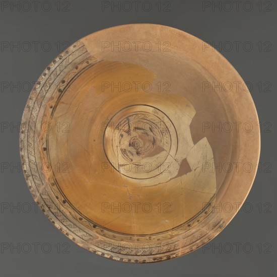 Deep plate with a spherically formed bowl. Horizontally protruding rim, wide, thicker at the edge. In the centre of the bottom is the image of a fish: body covered with scales, forked tail, two schematic dorsal fins. The fish is surrounded by three concentric circles; between the two closest to the center, a braided pattern runs to the right. On the edge, evenly spaced black dots are painted, connected by arced lines that form an oval space between them. Below the edge, a braided pattern runs to the right. In the highest part of the bowl, just below the break separating the rim from the bowl, a wavy line is painted with black dots in its bends. Pink clay, engobe ranging from yellow to light brown, decoration in black. 6th–7th century, Edfu; vessel. Height 12.1–14.2 cm, rim diameter 60 cm, foot diameter 19.1 cm. Maker: unknown Aswan workshop. Classification: bowls, painted Coptic vessels, figural decorations, Late Roman; braided and floral decorations., A large circular shallow bowl see...