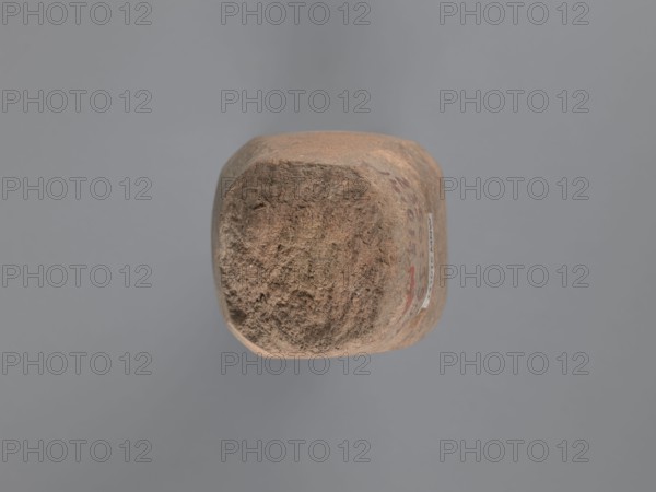 Pyramidal clay seal with a roughly square stamp surface and rounded corners. Shallow incisions at mid-height. Base engraved with a letter—interpreted as a geometric B or an M resting on the base— beside a small round recess. Upper part is impressed; chippings on the base. Visible kaolin clay, pink-beige. 6th–7th century. Origin: Edfu, southern Egypt. Seal, stamp; height 6.1 cm, width 5.5 cm. Geometric decorations, monogram/engraved decoration, stamp impressions. Maker/workshop: unknown South Egyptian workshop., A roughly cube-shaped object with rounded edges sits against a plain gray background; the object's surface shows a textured, weathered appearance with earthy tones of brown and tan, and there are traces of darker brown and faint red on one side.