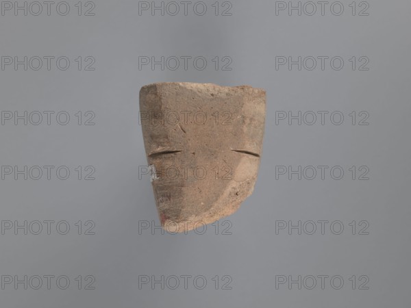 Seal, unknown South Egyptian workshop, pyramidal clay seal with a roughly square stamp surface and rounded corners. Shallow incisions at mid-height. On the base an engraved letter—possibly a geometric B or an M resting on the base—with a small round recess beside it. Upper part is impressed; chipping on the base. Visible kaolin clay, pink-beige. 6th–7th century; Edfu. Seal/stamp. Height 6.1 cm, width 5.5 cm., A weathered, fragmentary brown clay object with a roughly rectangular top and tapered lower edge suspended against a gray background, showing two shallow horizontal grooves on its front surface that suggest simple facial features and a rough textured surface with small pits and worn edges, with visible areas of darker brown and lighter tan variations and a small patch of white on one side.