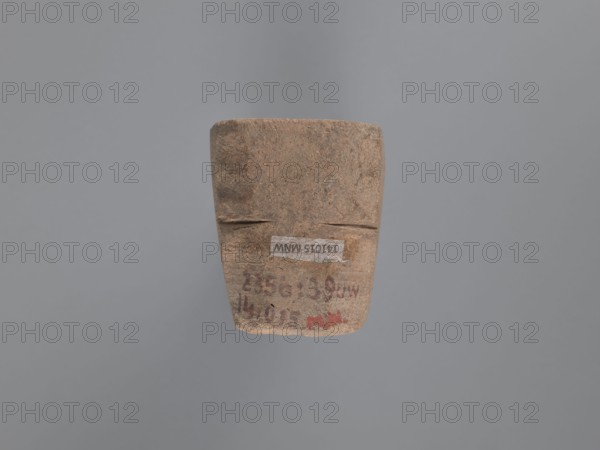 Seal, unknown South Egyptian workshop, pyramidal clay seal with a roughly square stamp surface and rounded corners. Shallow incisions at mid-height. On the base is an engraved letter—either a geometric B or an M resting on the base—next to a small round recess. Upper part is impressed; chippings on the base. Visible kaolin clay, pink-beige. 6th–7th century, Edfu; seal/stamp. Height 6.1 cm, width 5.5 cm., A roughly rectangular, weathered object with an uneven surface floats against a plain gray background; the object appears mostly brown with variations of tan and darker brown mottling, and a few faint areas of reddish discoloration.