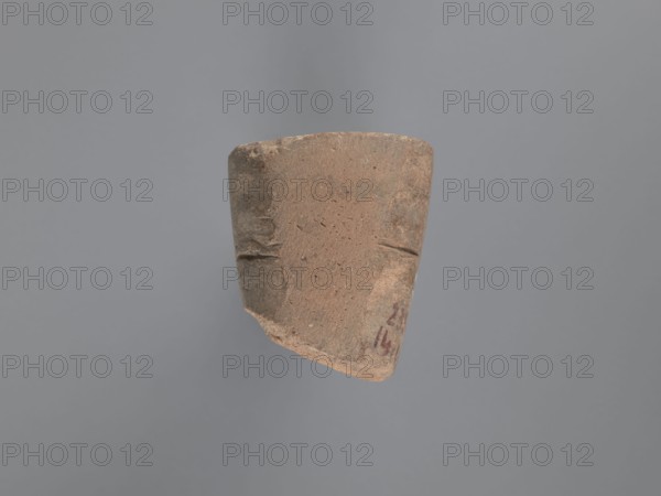 Seal, unknown South Egyptian workshop; pyramidal clay seal with a roughly square stamp surface, rounded corners; shallow incisions at mid-height; on the base an engraved letter possibly a geometrical B or an M resting on the base with a small round recess beside it; upper part is impressed, chippings on the base; visible kaolin clay, pink-beige; 6th–7th century; Edfu; seals, geometric decorations, monograms, engraved decorations, stamp impressions, stamps; seal/stamp; height 6.1 cm, width 5.5 cm, A roughly trapezoidal fragment of pottery floats against a neutral background. The fragment has a coarse, textured surface in shades of brown and tan with subtle darker brown areas and small speckles. The edges are worn and irregular, with a chipped corner showing a slightly lighter interior. There are a few shallow incised lines on the fragment’s face and faint darker smudges near one edge. The background appears as a smooth expanse of muted gray.
