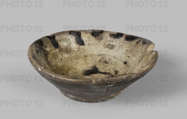 Bowl with a flat bottom, Islamic art, Arabic culture, Fatimid dynasty 909–1171. Arabic pottery, Egyptian pottery, household pottery for everyday life; fajumi vessel type, bowl with low walls and straight rim. Decoration: streaks of manganese pigment flowing from the rim edge, zigzag motif in the mirror referring to lettering; dark engobe on the reverse. 10th–12th century, Egypt, Edfu. Vessel; height 4.1 cm, diameter 12.0 cm., A shallow ceramic bowl with an irregular rim showing signs of wear, glazed interior with mottled beige and off-white tones and several dark brown to black blotches, outer surface in darker brown and gray shades with lighter worn spots, a chipped area on the rim and small surface cracks, all set against a plain light gray background.