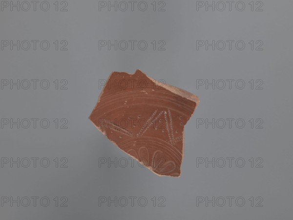 Plate fragment, unknown Aswan workshop site type / place of origin, plates, tableware, Aswan pottery, Egyptian pottery, Egyptian sigillata, Coptic vessels, Coptic pottery, impressed decorations, stamped decorations, stamps, kaolin clay. Fragment of the bottom of a plate with a ring-shaped foot. Pink kaolin ceramic mass Gempeler 1A. The surfaces are polished and covered with a reddish-brown engobe. The vessel is decorated internally, on the bottom of the bowl, using pressing and engraving techniques. These create a composition whose central element is a rosette impressed with almond-shaped stamps forming a pattern. It is surrounded by five concentrically engraved grooves. The band surrounding them consists of a double-course zigzag, made with an impressed roulette. The fields between the zigzag arms are filled with rhomboidal impressions of a stylus . The entire composition is enclosed by eight concentrically engraved grooves. 5th–6th century, unknown type of site / place of discovery, ...