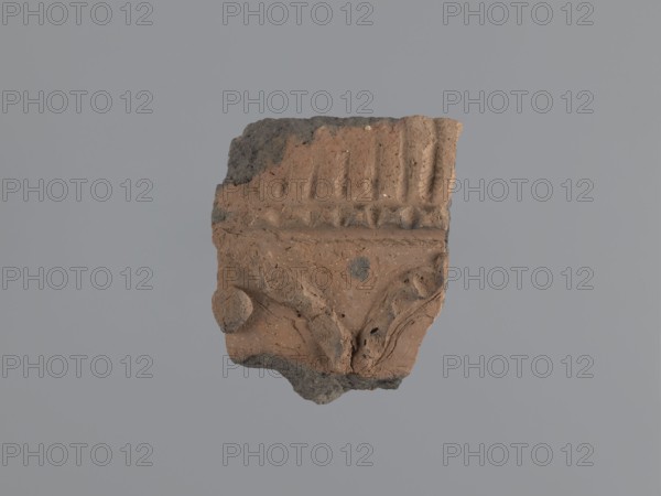 Fragment of a utility vessel Utility Ware. Made of type C Nile clay, highly porous with organic admixtures such as chaff and gold mica flakes. Thick fracture, dark gray Unworked surface exterior and interior brown, Engraved and plastic decoration, hand-applied. A horizontal half-cylinder, plastically modeled with recesses made with fingers or a round tool, separates the upper from the lower part. At the top is a row of parallel recesses additionally decorated with a knurl. In the lower section are two partially preserved finger-shaped arcades made of glued-on clay rolls; a knob made from a clay disc is glued under one arcade. Wavy engraved lines run along the arcades. Above the arcades is a horizontal row of diagonal recesses—remains of the imprint of the rope used to tie the vessel during drying. 6th–7th century, Tell Atrib; vessel, height 14 cm, width 11 cm, thickness 2 cm., A fragment of a textured, irregularly shaped object with earthy reddish-brown and darker gray areas, showing a...