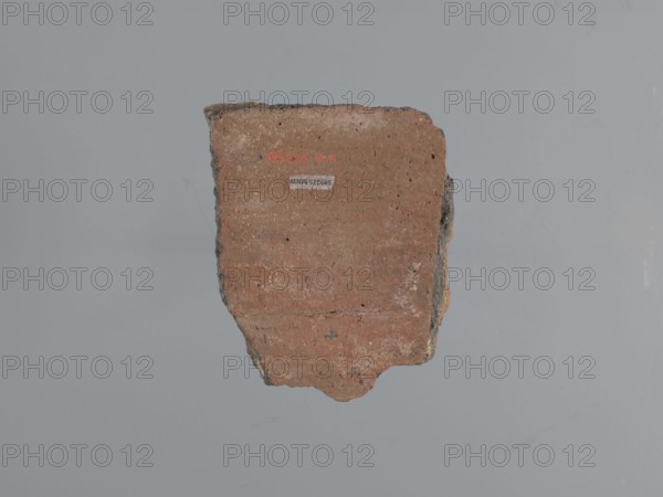 Fragment of a utility vessel. Made of type C Nile clay, highly porous with organic admixtures chaff and gold mica flakes. Thick fracture, dark gray Unworked surface exterior and interior brown, Engraved and plastic decoration, hand-applied. A horizontal half-cylinder, plastically modeled with recesses made with fingers or a round tool, separates the upper from the lower part. Top: a row of parallel recesses additionally decorated with a knurl. Lower: two partially preserved finger-shaped arcades made of glued-on clay rolls; a knob made from a clay disc is glued under one arcade. Wavy engraved lines run along the arcades. Above the arcades is a horizontal row of diagonal recesses—remains of the rope imprint used to tie the vessel during drying. 6th–7th century. Tell Atrib. Vessel; height 14 cm, width 11 cm, thickness 2 cm., A single irregular fragment of a hard material with a rough texture is centered against a plain background. The fragment shows a flat face with visible horizontal st...