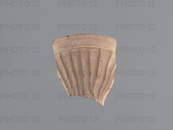 Fragment of a conical bowl or possibly a large funnel, tabletop or kitchen vessel. Egyptian workshop; bowls with engraved decorations, Coptic vessels. Made of type B Nile clay, compact with a small amount of organic admixtures and particles of golden mica. Fracture shows a dusty pink outer and dark grey core 7.5R5/4-10YR4/1-7.5R5/4. Outer surface finished with pink-tinged whitewash 5YR8/3; inner surface unfinished light reddish-brown, 2.5YR6/3. Straight spout with rounded edge. Outer surface below the rim decorated with vertical incised bands made with a stick or reed; incisions converge toward the bottom. Fragment was likely reused, as two side fractures are covered with a thin layer of gypsum mortar; similar substance found on the inner surface near one fracture. 7th–8th century, Tell Atrib. Vessel: height 14.7 cm; width 13.1 cm; thickness 8 mm; rim diameter 21 cm., A fragment of a ridged ceramic or terracotta object with vertical fluted grooves and a rounded top edge sits centered a...