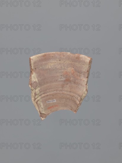 Fragment of a conical bowl or possibly a large funnel, tabletop or kitchen vessel. Made in an Egyptian workshop of type B Nile clay, compact with slight organic admixture and particles of golden mica. Fracture shows a dusty pink and dark grey core 7.5R5/4–10YR4/1–7.5R5/4. Outer surface finished with pink-tinged whitewash 5YR8/3; inner surface unfinished light reddish-brown, 2.5YR6/3. Straight spout with a rounded edge. Outer surface below the rim decorated with vertical incised bands made with a stick or reed, the incisions converging toward the bottom. Likely reused, as two side fractures are covered with a thin layer of gypsum mortar and a similar substance is present on the inner surface near one fracture. 7th–8th century, Tell Atrib. Vessel: height 14.7 cm, width 13.1 cm, wall thickness 8 mm, rim diameter 21 cm., A broken fragment of a rounded ceramic object with curved ridges and an uneven edge sits against a flat gray background; the fragment shows a worn, earthy surface with sha...
