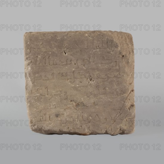 Fragment of a stele with an inscription in Arabic, unknown creator/scribe, excavated in Faras Sudan, Islamic art, Arabic inscriptions in Kufi script containing a fragment of the Quran 67:1; probably a funerary stele. 2nd half of the 11th–1st half of the 14th century. Origin: Faras, Sudan. Stele; height 32.0 cm, width 34.0 cm, depth 12.0 cm. The surface of the block is smoothed., A roughly square, flat stone slab with an uneven, worn surface and rounded edges, shown against a neutral light gray background; the slab is primarily shades of brown and tan with subtle variations and darker brown spots, faint incised lines and shallow depressions visible across its face, and a few small chips and a crack along one side; the overall texture appears rough and weathered.