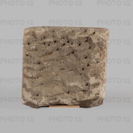 Fragment of a stele with an inscription in Arabic, unknown creator scribe-copyist, excavations, Islamic art, Arabic culture, inscriptions, Arabic inscriptions, Kufi script, Quran, Arabic language, Nubia, Nubian crafts. The surface of the block is smoothed; the inscription in Kufi script contains a fragment of the Quran 67:1. It is probably a funerary stele. 2nd half of the 11th–1st half of the 14th century. Origin: Faras, Sudan. Stele; height 32.0 cm, width 34.0 cm, depth 12.0 cm., A roughly rectangular block with a rough, pitted surface sits centered against a uniform light gray background, the block showing a textured, weathered appearance in shades of brown, beige, tan, and hints of darker brown in small pits and indentations; the top and sides display irregular grooves and a few lighter patches, while a thin flat support in a light wood color is visible under the block at the front.