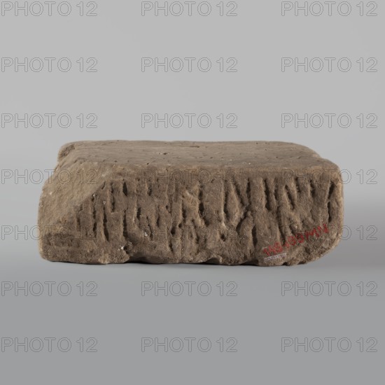 Fragment of a stele with an inscription in Arabic, /scribe, excavations, Islamic art, Arabic culture, inscriptions, Arabic inscriptions, Kufi script, Quran, Arabic language, Nubia, Nubian crafts. The surface of the block is smoothed; the inscription in Kufi script contains a fragment of Quran 67:1. It is probably a funerary stele. 2nd half of the 11th–1st half of the 14th century. Sudan, Faras. Stele; height 32.0 cm, width 34.0 cm, depth 12.0 cm., A rectangular block with rounded corners sits on a flat surface; the block appears textured and rough with irregular vertical grooves and small pits on its front face, and a flatter, slightly uneven top surface. The visible colors are shades of brown on the block, with a few small lighter spots, and a neutral gray background and surface.