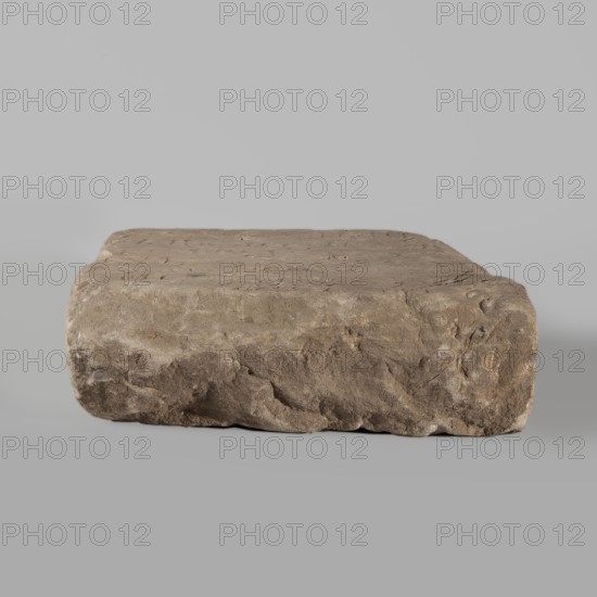 Fragment of a stele with an inscription in Arabic, unknown creator/scribe, excavations, Islamic art, Arabic culture, inscriptions, Arabic inscriptions, Kufi script, Quran, Arabic language, Nubia, Nubian crafts. The surface of the block is smoothed; the inscription in Kufi contains a fragment of Quran 67:1. It is probably a funerary stele. 2nd half of the 11th–1st half of the 14th century. Sudan, Faras. Stele; height 32.0 cm, width 34.0 cm, depth 12.0 cm., A rectangular block of rough stone with an uneven, weathered surface sits centered against a smooth background; the stone shows shades of brown, tan, beige, gray, and hints of darker brown in crevices, with textured irregular edges and small pits on the top and sides.