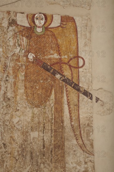 Wall painting: Archangel Gabriel with a sword, unknown artist. The Archangel is depicted standing frontally with feet slightly apart, holding a sheathed sword: his right hand grasps the crossguard while his left supports the sword halfway along its length. His wings droop downwards and are slightly curled at the tips. The Archangel has a high forehead, straight nose, almond-shaped eyes, small mouth; hair tied back with two locks over the forehead. He wears a long white tunic with rows of black potamoi and decorative cuffs, a yellow chlamys decorated with a diagonal red check fastened with a fibula on the right shoulder, and a green maniakion superimposed on the chlamys. His head is topped with a stemma-type crown partially destroyed due to damage to the top layer of plaster and surrounded by a yellow halo. The silhouette is significantly elongated, with hands and face small in relation to the figure’s height, producing a slightly Mannerist appearance emphasized by a facial grimace: str...