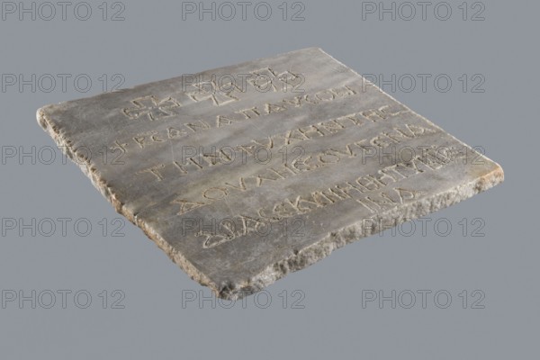 Epitaph of Gennadii. Inscriptions, Greek inscriptions, tombstone in fine-crystalline blue-gray marble. The lower edge of the slab is chipped; the preserved part is cracked transversely halfway up. In the upper part, above the inscription, three Maltese crosses are arranged symmetrically. Inscription in five lines, rounded letters, 1.5–2.0 cm high, average line spacing 2.5 cm. 6th–7th century, Constantinople, Byzantine Empire 476–1453. Greek inscription, epitaph. Height 29.5 cm, width 29.5 cm, depth 2.0 cm., A flat rectangular stone slab rests against a neutral gray background, showing a worn and uneven surface with a rough chipped edge. The slab is primarily gray with areas of beige and light brown along the edges and within shallow grooves. The top surface displays a few carved linear grooves and a group of shallow incised marks arranged in rows, and there are a few cross-shaped carvings near one edge. The stone shows subtle variations in tone from darker gray patches to lighter gray ...
