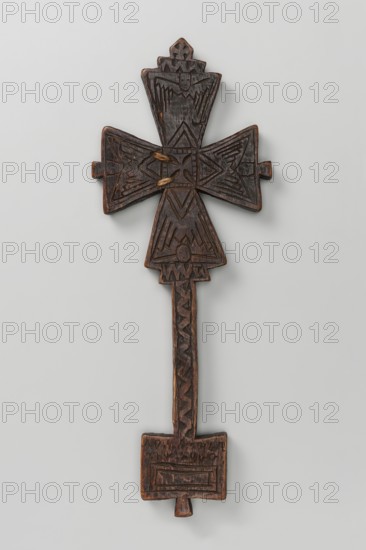Hand-made Ethiopian cross, 19th century. The arms are triangular with central projections on each arm, flowing into a handle in the form of an elongated rectangle topped by a rectangular plaque with a central lower projection. Decoration on both sides. Side A: Center has a small equal-armed cross with widening arms enclosed in a square decorative frame; triangles marked with double engraved lines appear on each side of the frame. The fields of the arms show schematic representations of four winged cherubim/seraph heads. The upper arm projection is decorated with a horizontal zigzag stripe and a cross. The handle has a zigzag stripe. The tablet features an Ethiopian inscription with a rectangle marked by a double line below it. Side B: Center has a small cross composed of five rectangles, from which four rhombuses filled with a quatrefoil motif emerge. The upper arm shows a schematic winged head cherub/seraph. The handle is undecorated. The tablet is decorated with a rectangular frame. ...