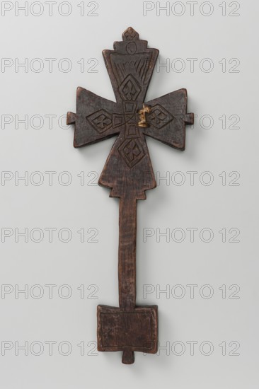 Hand-made Ethiopian cross. The arms are triangular with projections in the center of each arm, flowing into a handle in the form of an elongated rectangle, topped with a rectangular plaque with a projection in the center of the lower side. Decoration on both sides. Side A: In the center, a small equal-armed cross with widening arms, enclosed in a square decorative frame. On each side of the frame, a triangle marked with a double engraved line. In the fields of the arms are schematic representations of four winged cherubim or seraph heads. The projection of the upper arm is decorated with a horizontal zigzag stripe and a cross. The handle has a zigzag stripe. The tablet features an Ethiopian inscription, with a rectangle marked with a double line below it. Side B: In the center, a small cross composed of five rectangles, from which four rhombuses filled with a quatrefoil motif emerge. The upper arm features a schematic representation of a winged head—a cherub or seraph. The handle is un...