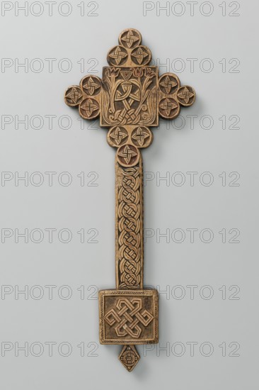 Hand cross, Ethiopian workshop. The arms of the cross are each formed by three stacked discs adjacent to one side of the square forming the central part of the cross. The handle, in the shape of an elongated rectangle, merges into a rectangular plaque with a rhomboid projection at the lower edge. Decoration on both sides. Side A: Within the square is a carved motif of a cross made of braid. From the base of the cross grow stylized flowers resembling tulips—one on the left and two on the right. Within the discs is a quatrefoil motif made of braid. The handle is decorated with braid. The plaque is decorated with braid framed in a zigzag strip. The inset is decorated with a braid. Side B: Square decorated with a crystal ornament; discs divided diagonally into eight wedges. The handle is decorated with a braid. The plate is decorated with a braided quatrefoil motif. The inset is decorated with a braided motif. 20th century, Ethiopia. Cross. Height 27.5 cm; width 10.5 cm., A carved wooden o...