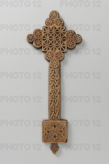 Hand cross, Ethiopian workshop. The arms of the cross are each formed by three stacked discs adjacent to one side of the square forming the central part of the cross. The handle, in the shape of an elongated rectangle, merges into a rectangular plaque with a rhomboid projection at the lower edge. Decoration on both sides. Side A: Within the square is a carved motif of a cross made of braid. From the base of the cross grow stylized flowers resembling tulips—one on the left and two on the right. Within the discs is a quatrefoil motif made of braid. The handle is decorated with braid. The plaque is decorated with braid framed in a zigzag strip. The inset is decorated with a braid. Side B: Square decorated with a crystal ornament; discs divided diagonally into eight wedges. The handle is decorated with a braid. The plate is decorated with a braided quatrefoil motif. The inset is decorated with a braided motif. 20th century, Ethiopia. Cross. Height 27.5 cm; width 10.5 cm., A carved wooden o...