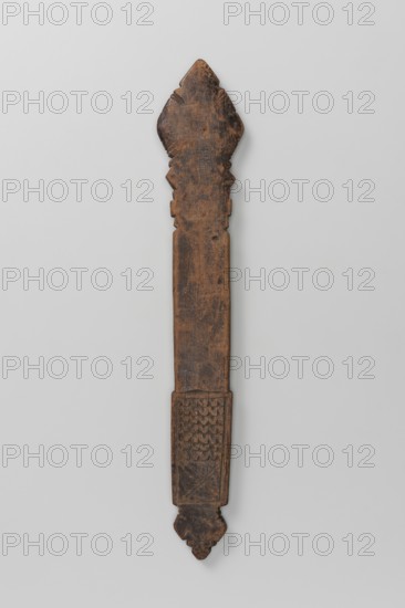 Hand cross, Ethiopian workshop. Diamond-shaped arms, wide grip almost same width as the arms, merging into a poorly defined plaque in the shape of an elongated rectangle topped with a trefoil inset. Bilateral decoration almost identical on sides A and B, limited to simple incisions at the edges of the cross arms and horizontal zigzag stripes and a double X on the plaque. 16th–20th century. Ethiopia. Cross; height 28 cm, width 5 cm., A long narrow wooden object with a weathered brown surface, carved with a pointed ornament at the top and a decorated rectangular section near the bottom; the top and bottom ends feature symmetrical carved shapes and the wood shows worn texture and darker brown variations against a pale gray background.