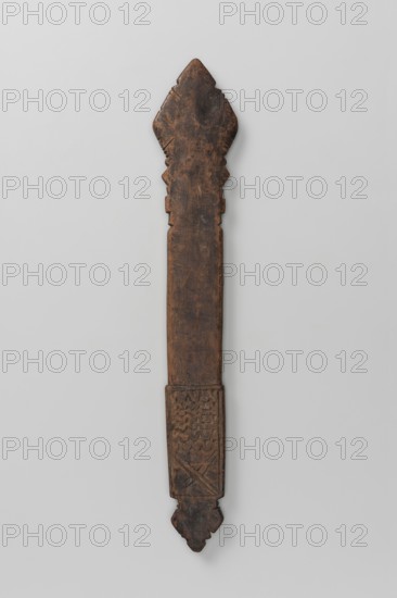 Hand cross, Diamond-shaped arms, wide grip almost same width as the arms, merging into a poorly defined plaque in shape of an elongated rectangle topped with a trefoil inset. Bilateral decoration, almost identical on sides A and B, limited to simple incisions at the edges of the cross arms and horizontal zigzag stripes and a double X on the plaque. 16th–20th century, Ethiopia. Metal cross, height 28 cm, width 5 cm., A long, narrow carved wooden object with a dark brown, weathered surface against a light gray background; the top end is shaped with a pointed, scalloped outline and the bottom end tapers to a decorative, rounded tip with carved geometric motifs near the lower section; the central shaft is relatively smooth and rectangular, showing natural wood grain and worn areas, and the lower portion features incised patterns with zigzag and cross-hatched textures.