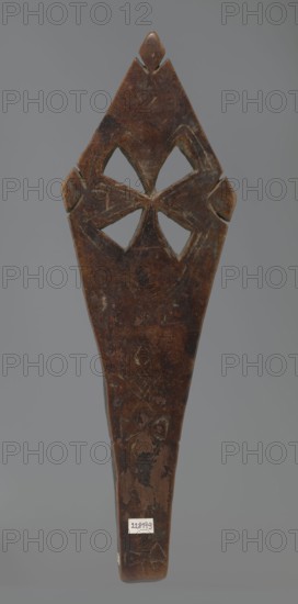 Hand cross fragment, Simple engraved decoration on the surface: four triangular cutouts arranged in the form of a cross; below the holes are two rather carelessly engraved crosses. The reverse is also decorated with a cross motif; decoration engraved with a thin, shallow line. Dark wood. 20th century, Ethiopia. Cross; height 26 cm, width 8 cm., A vertically oriented carved wooden object with a dark brown surface showing lighter worn areas and scratches, featuring a pointed top with a small raised diamond-shaped element and carved cutouts forming a symmetrical four-petaled motif near the upper portion, flanked by small protruding shapes on each side; the lower portion tapers into a handle-like shape with visible patches of lighter and darker brown wear and some faint carved line patterns, all set against a neutral gray background with a few small areas of lighter discoloration on the wood.