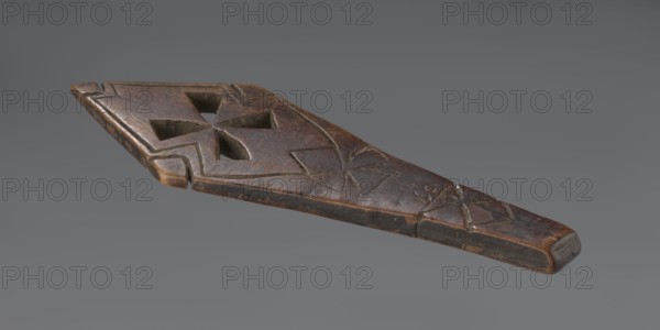 Hand cross - fragment. Simple engraved decoration on the surface; four triangular cutouts arranged in the form of a cross. Below the holes, two rather carelessly engraved crosses are visible. The reverse is also decorated with a cross motif. Decoration engraved with a thin, shallow line. Dark wood. 20th century, Ethiopia. Cross; height 26 cm, width 8 cm., An elongated carved wooden object with a tapered end and a wider opposite end, showing a carved geometric cutout near the wider end and shallow incised linear decorations along its surface; the wood surface displays worn and weathered areas with a patina ranging from dark brown to lighter brown and small patches of black, set against a plain grey background.